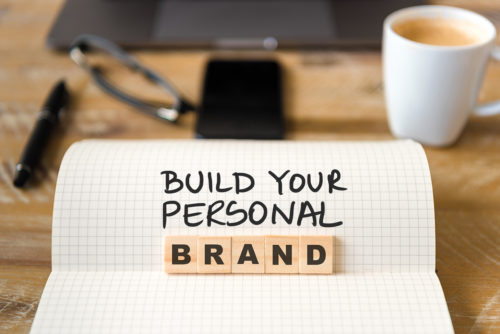 Closeup on notebook over vintage desk surface, front focus on wooden blocks with letters making Build Your Personal Brand text. Business concept image with office tools and coffee cup in background